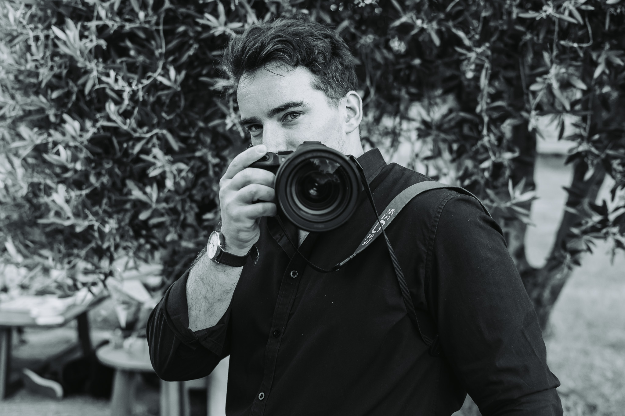 Black and white portrait of a photographer looking over a Canon camera among outdoor foliage