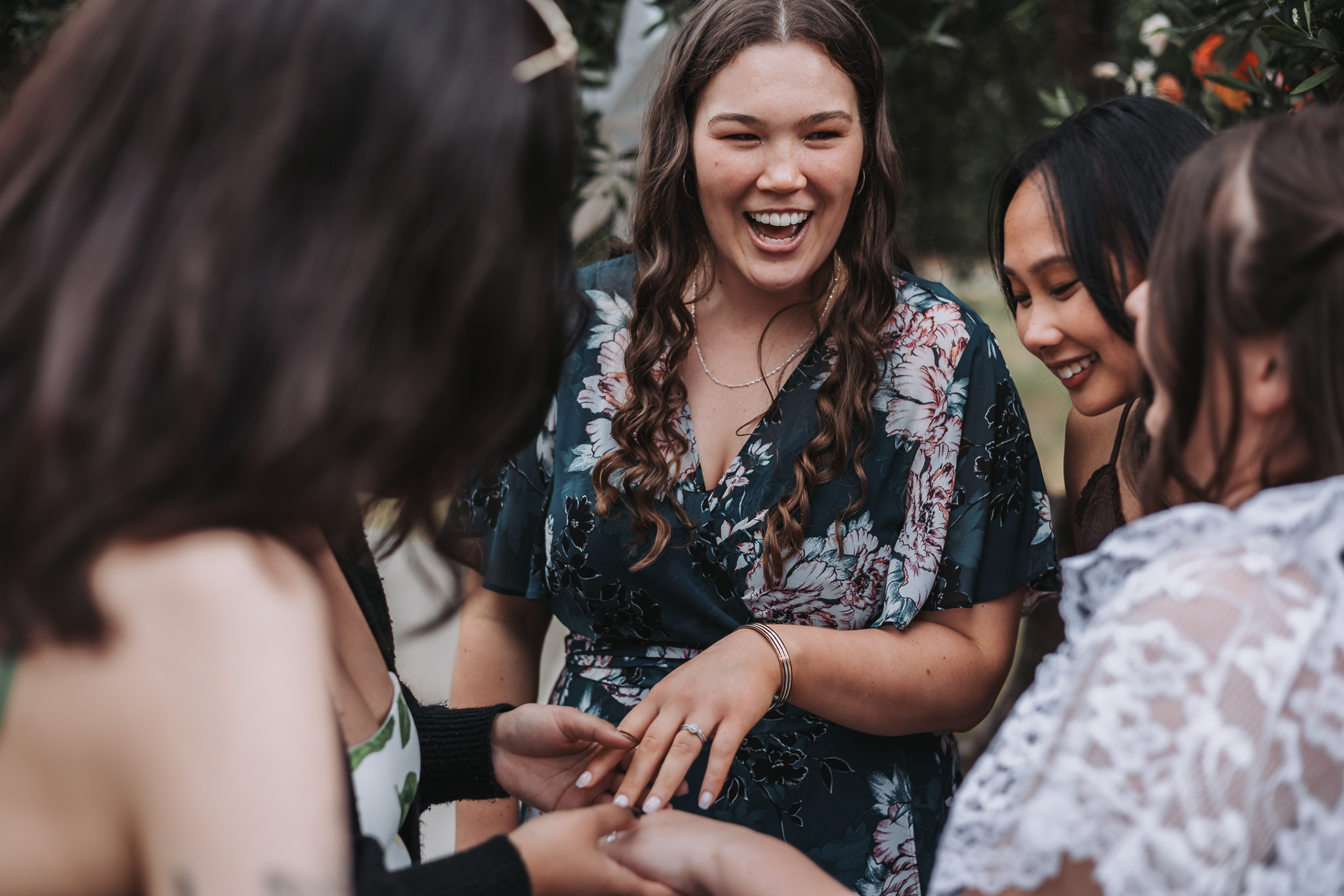 Woman laughing as friends gather around to admire her engagement ring in a garden setting