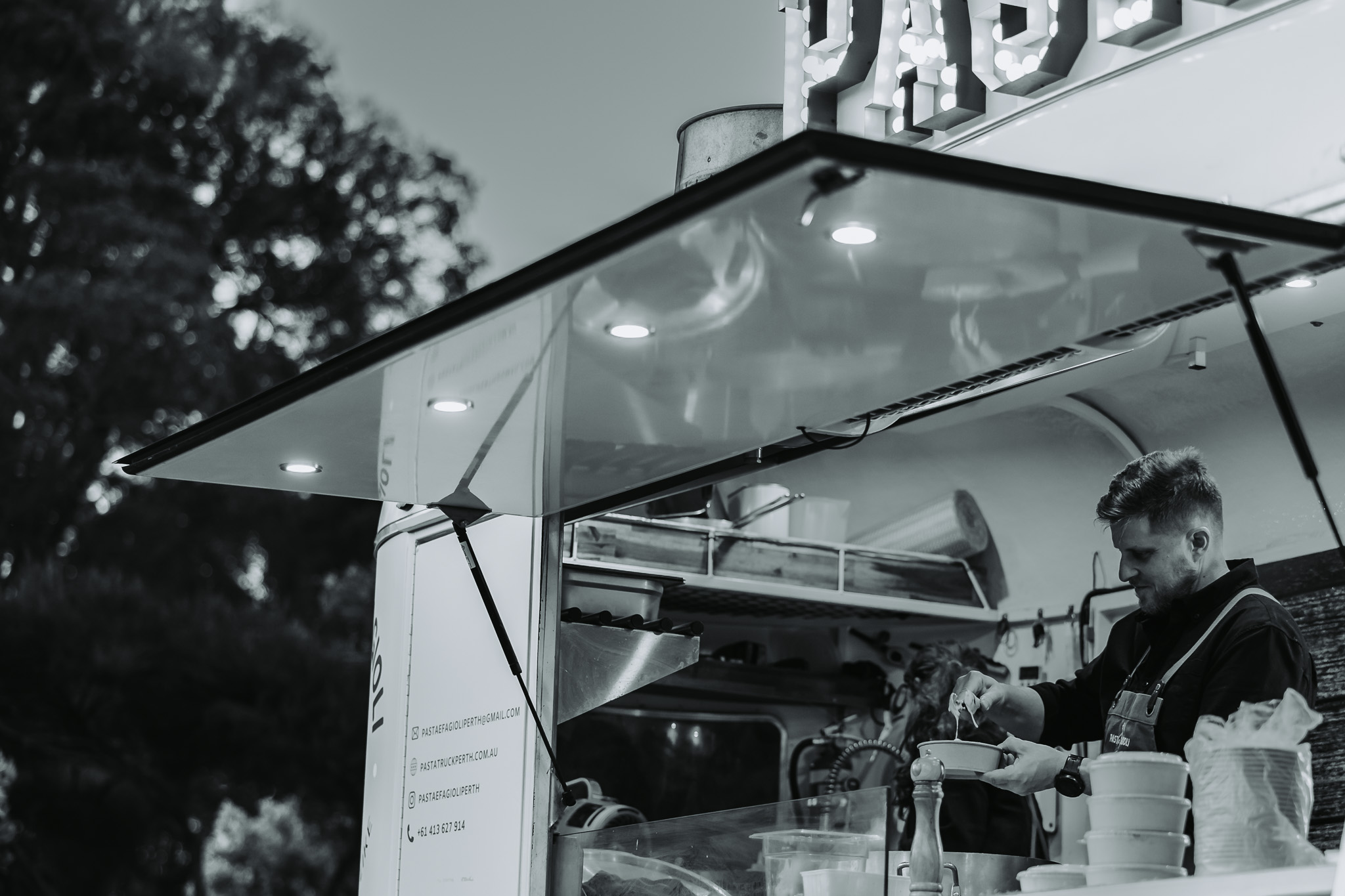 Black and white photo of a chef working from a lit pasta food truck at an evening Perth event
