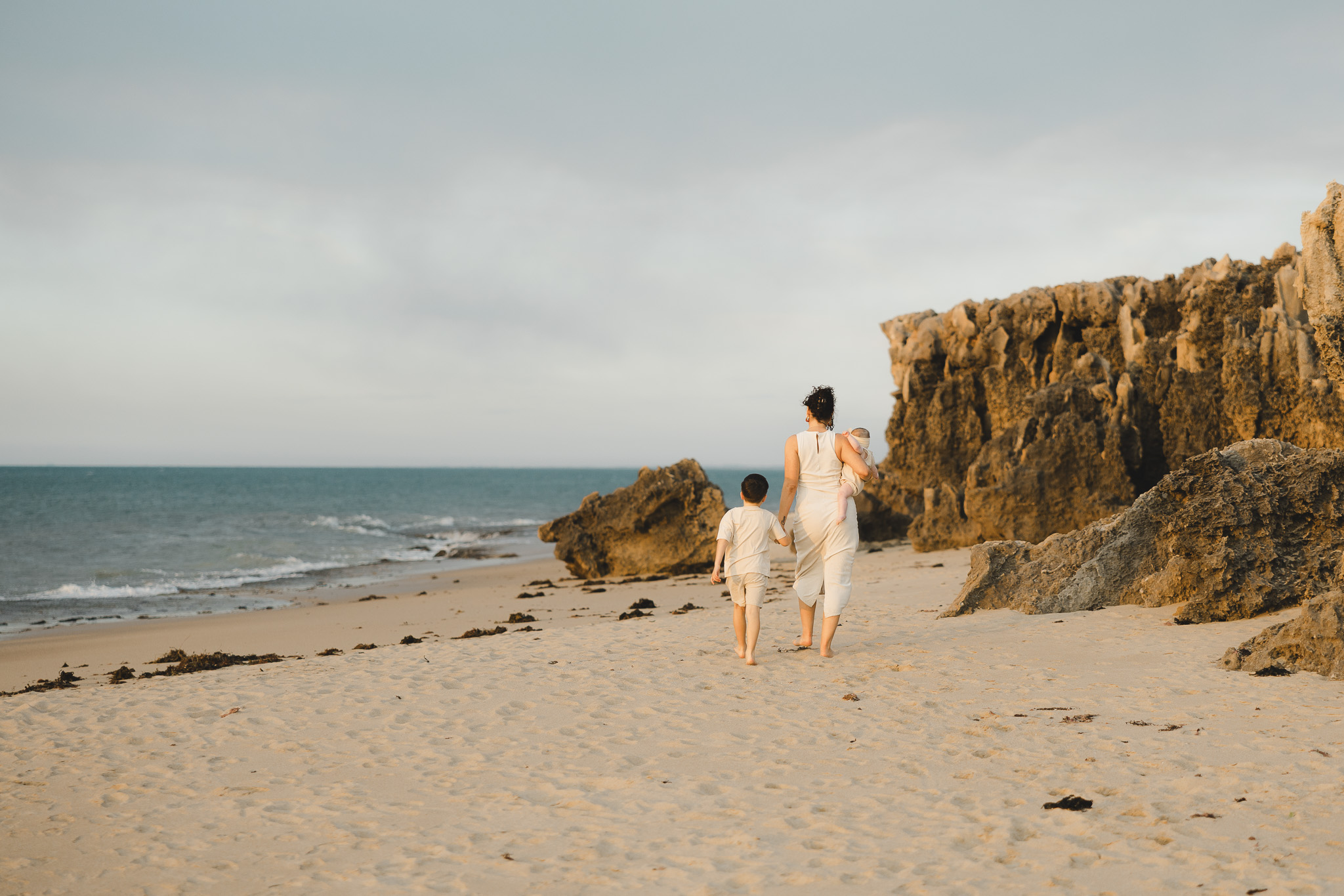 Mother carrying a baby while walking barefoot along a rocky Perth shoreline with her young son