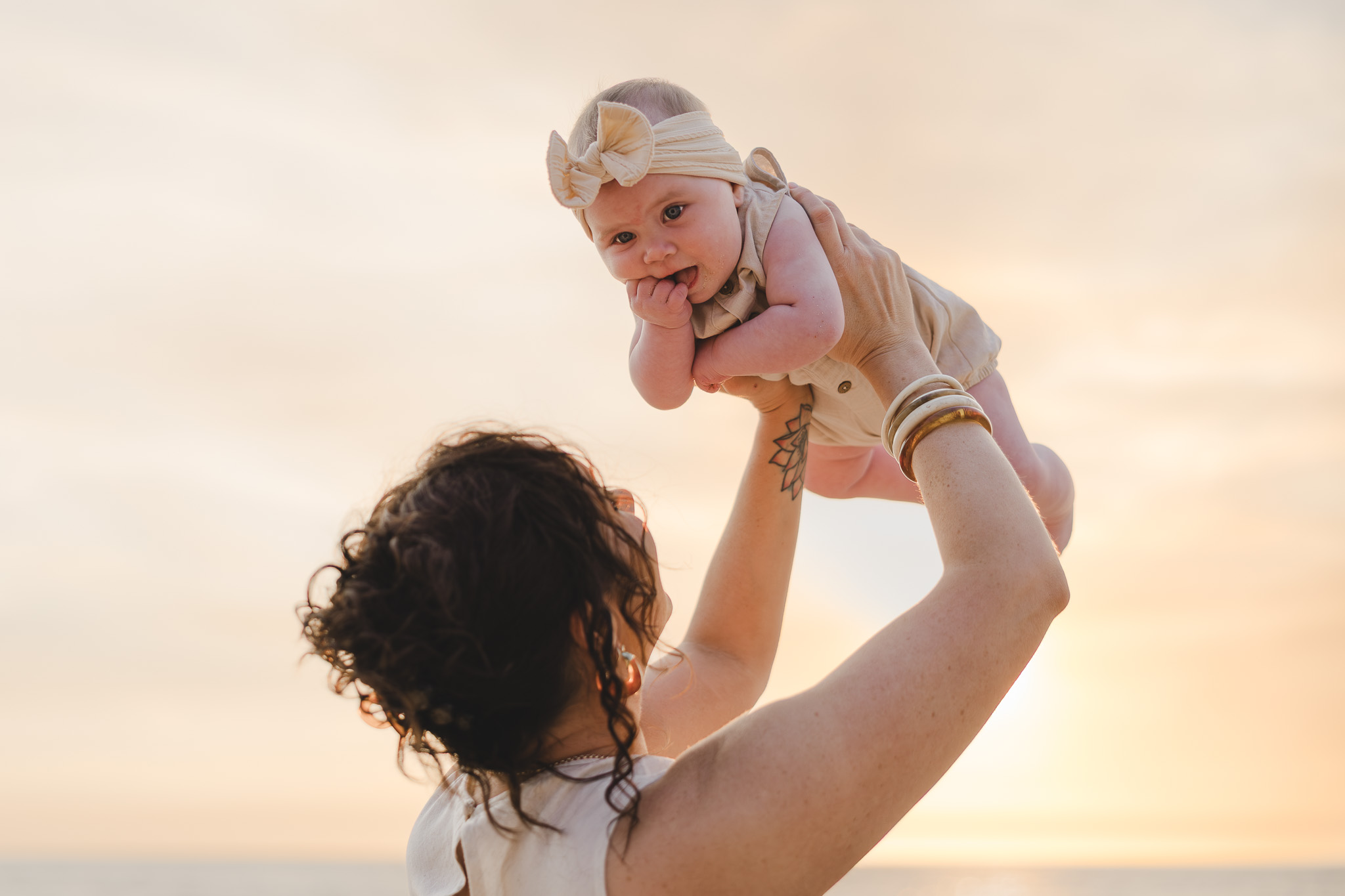 Mother lifting her baby into the air against a soft golden sunset sky