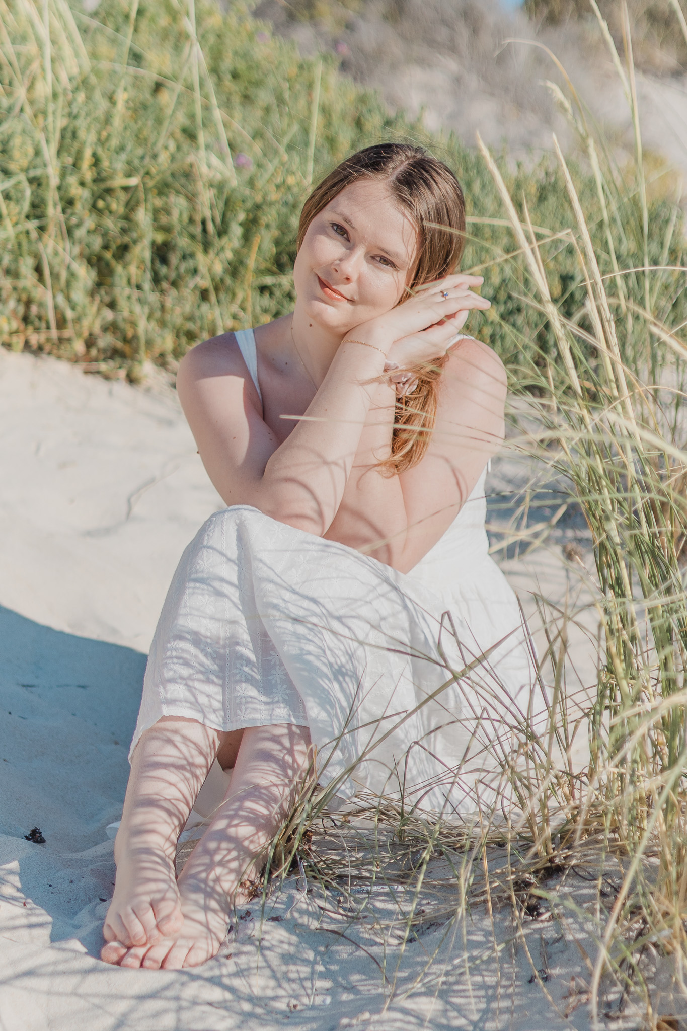 Woman in a white eyelet sundress sitting among beach grass dunes, smiling softly