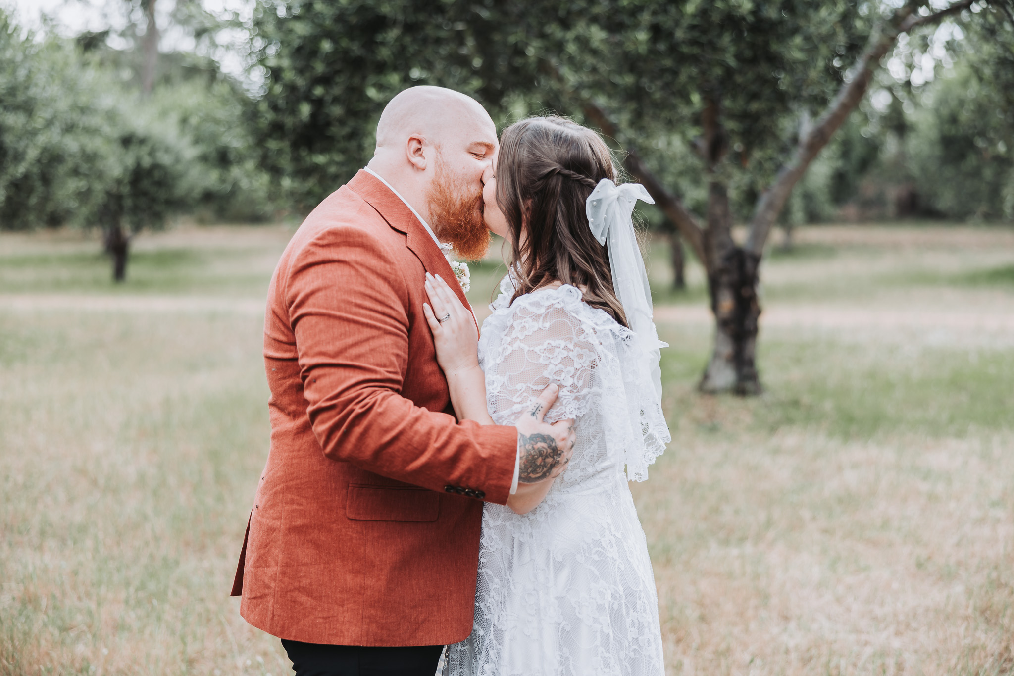 Bride and groom sharing a kiss outdoors — lace dress and terracotta blazer among olive trees