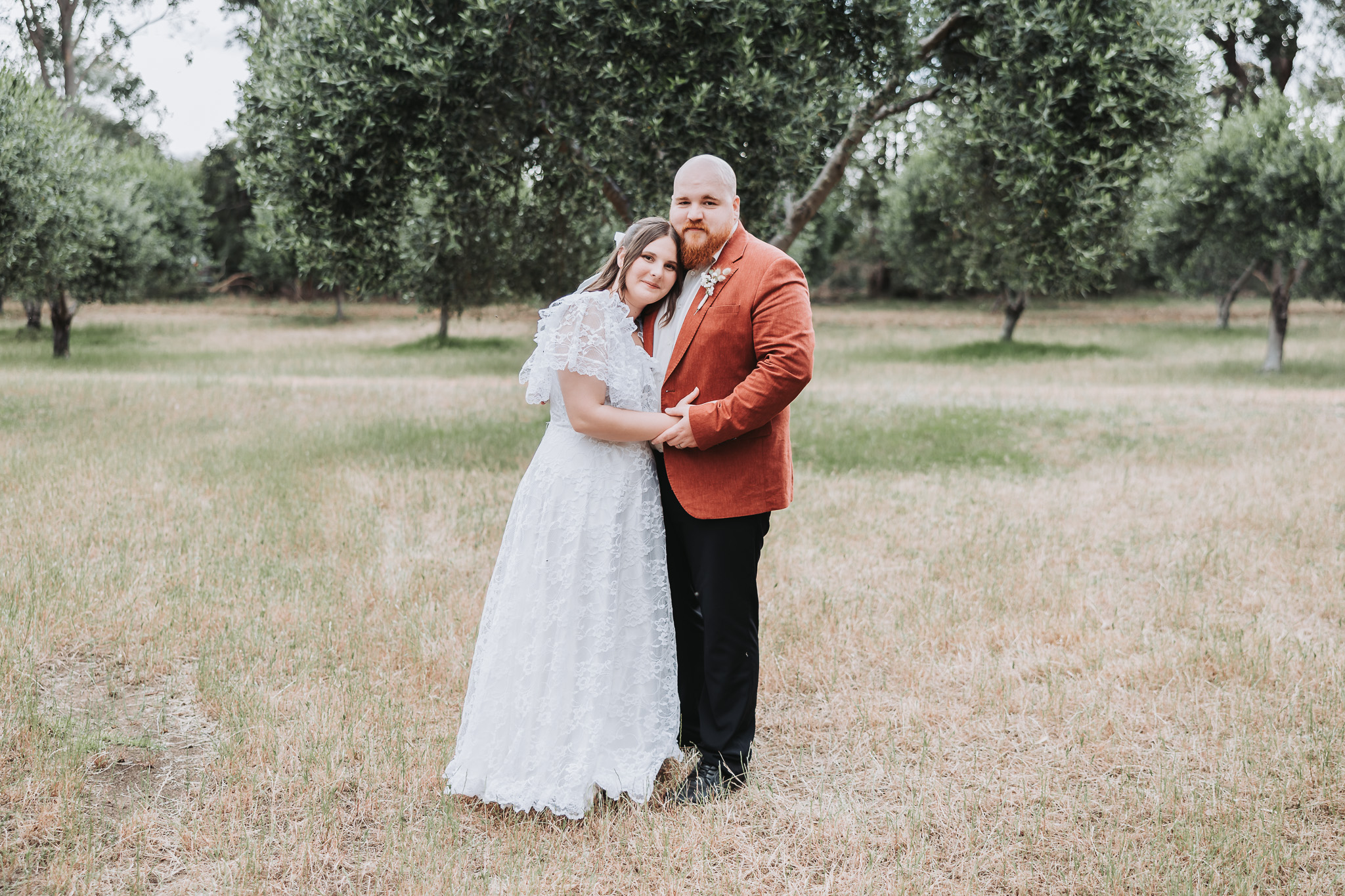 Bride in a lace wedding dress and groom in a terracotta blazer posing in an olive grove