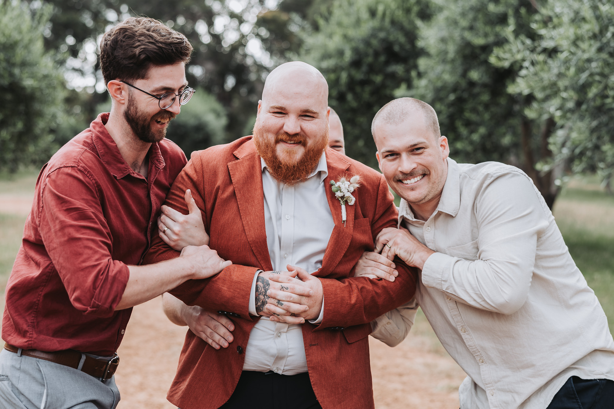 Groom in a terracotta blazer laughing with two friends in an outdoor orchard at a Perth wedding